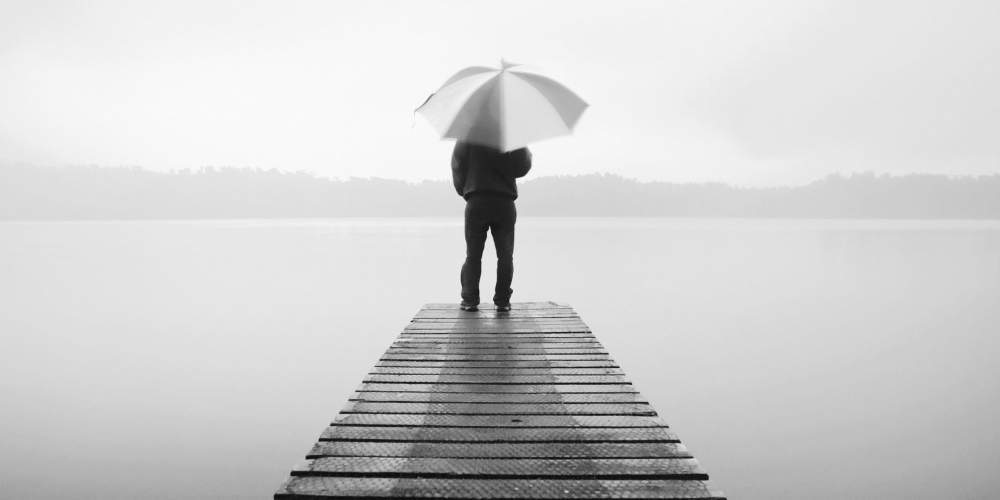 Auf diesen Bild ist ein Mann mit einem Regenschirm am Ende eines Stegs im Nieselregen zu sehen und symbolisiert eine Situation im Prozess zur Heilung der Existenzangst